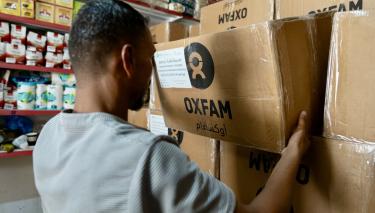 Verteilung von Nahrungsmittelpaketen mit haltbaren Lebensmitteln in Nord-Gaza. Ein Mann hält einen Karton mit Oxfam-Logo. Im Hintergrund sind Regale mit Nahrungsmitteln zu sehen.